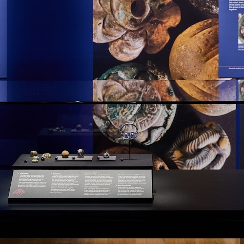 Museum exhibit featuring small ancient artifacts in a glass case with descriptive placards. Large artifact images are displayed behind.