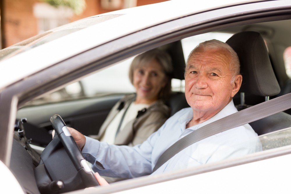 Older male driver sat in a vehicle smiling with a older female in the passenger seat also smiling