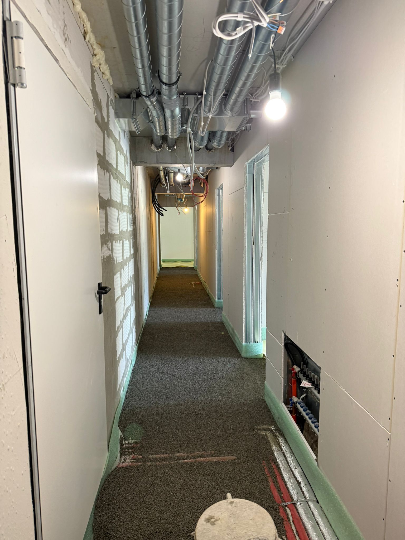 Schmaler Flur mit Türen auf beiden Seiten, unverputzten Wänden und freiliegenden Rohren und Kabeln an der Decke.
