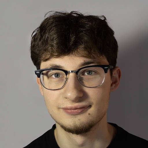 A young man with glasses and a denim shirt sits confidently against a blue wall. Art and cacti decorate the space, conveying a creative vibe.