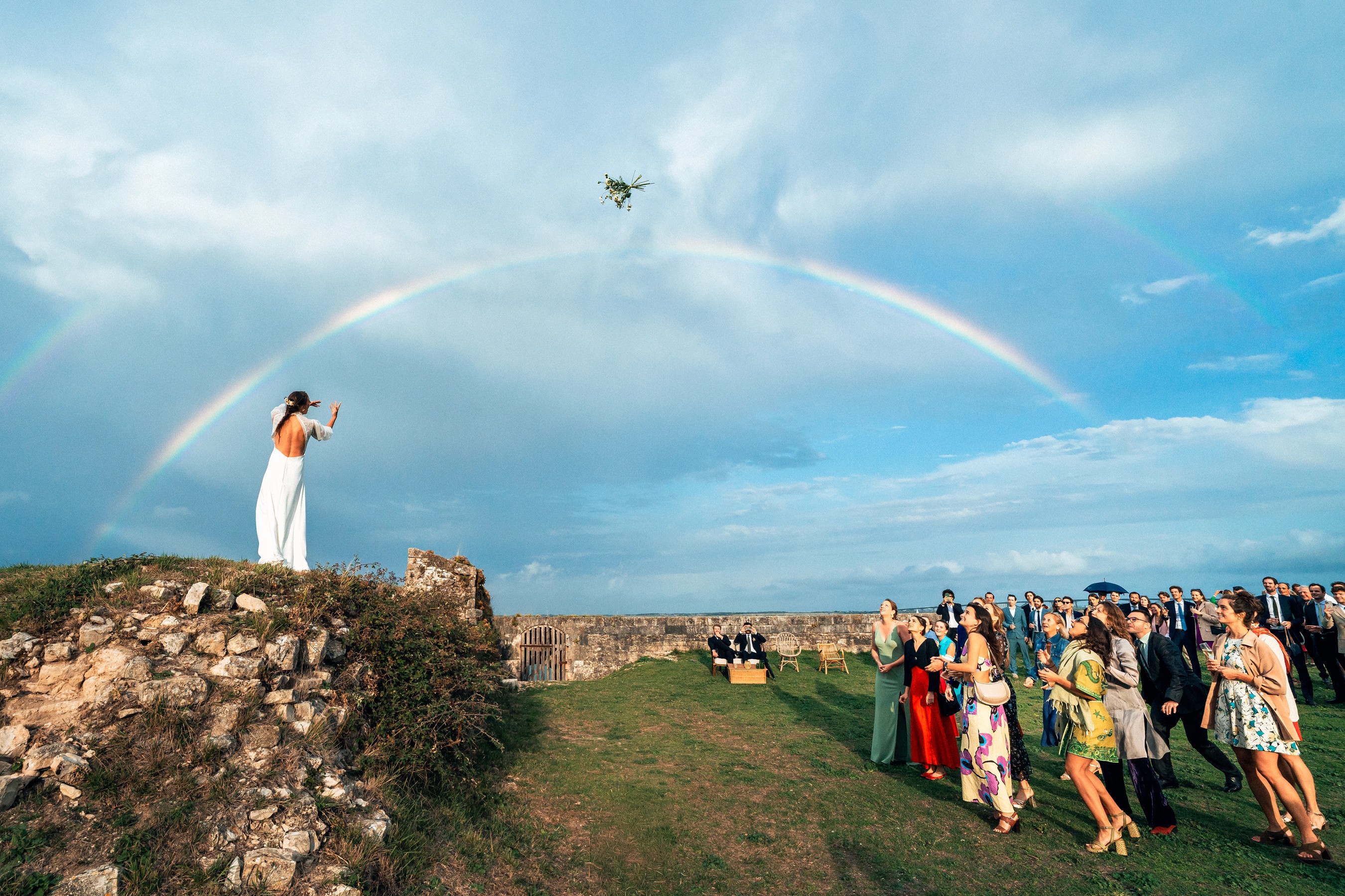 Photographe de mariage au Pays basque, bouquet lancé dans le ciel sous un arc-en-ciel, symbolisant une vision humaine, intense et poétique du mariage.