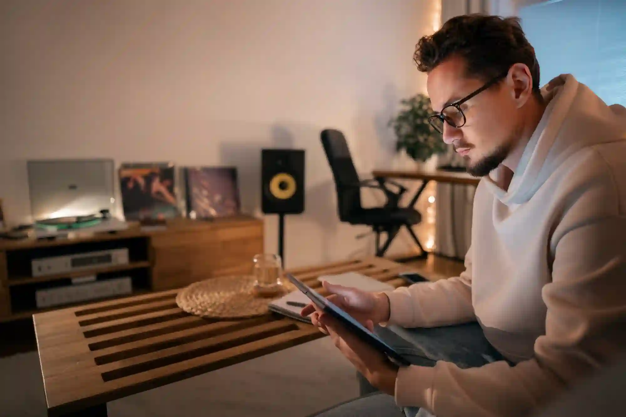 A man in a hoodie sits in a dimly lit room, focused on his tablet with music equipment behind.