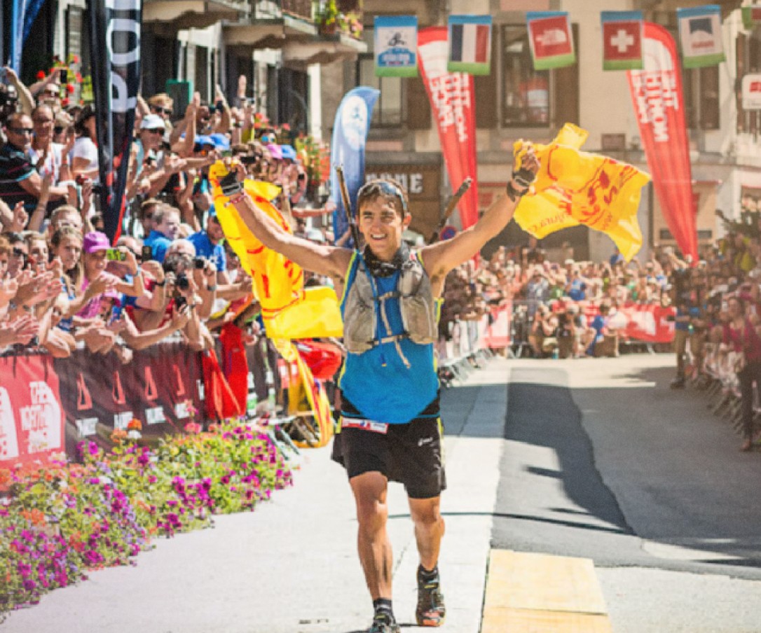 Un coureur franchit la ligne d'arrivée d'une course en montagne entourée de spectateurs enthousiastes, avec des bannières aux couleurs vives et un sourire victorieux.