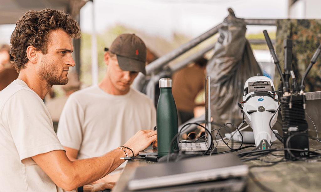 Two individuals work at a cluttered outdoor workstation with laptops, a reusable water bottle, and various electronic devices, highlighting a focus on technology and mobile communication.