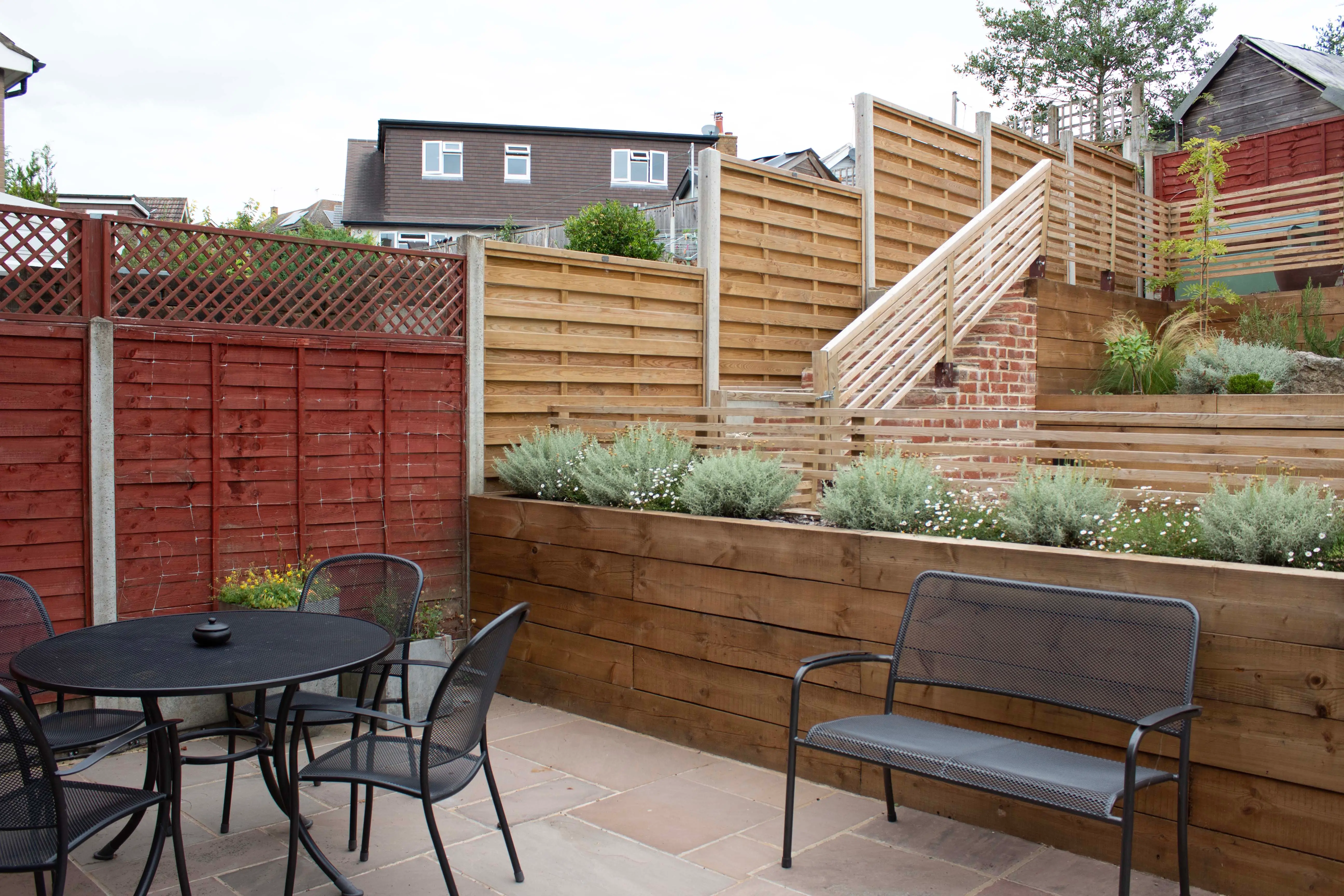 A patio area with a table and chairs, surrounded by wooden fencing and a landscaped garden with stairs in the background.