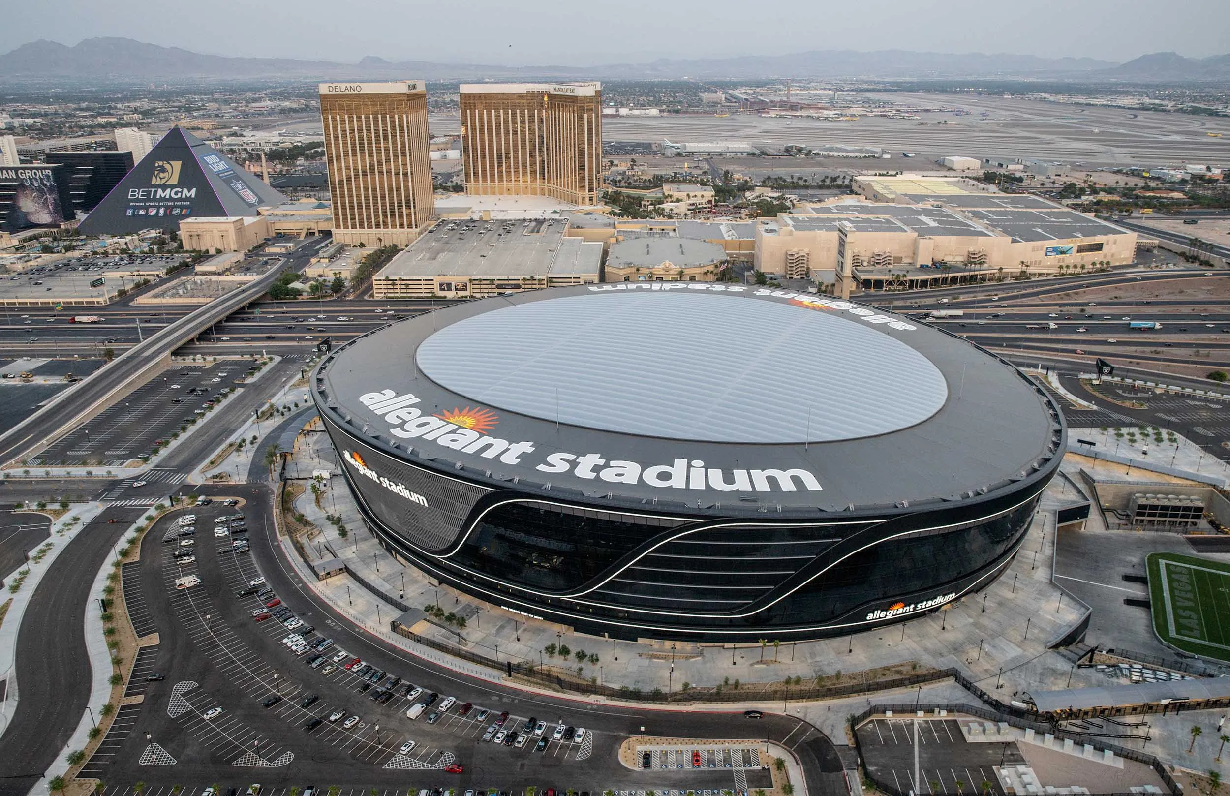 Aerial shot of Allegiant Stadium surrounded by Las Vegas hotels and highways, with the Luxor and Mandalay Bay visible in the background. Captures the stadium’s massive scale and prime location on the Las Vegas Strip.