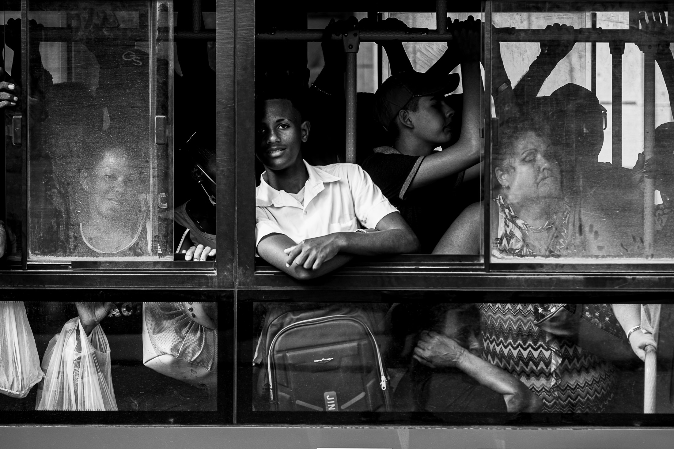 Crowded bus interior with passengers pressed together; a young man leans on the window frame while others stand and sit behind the glass.