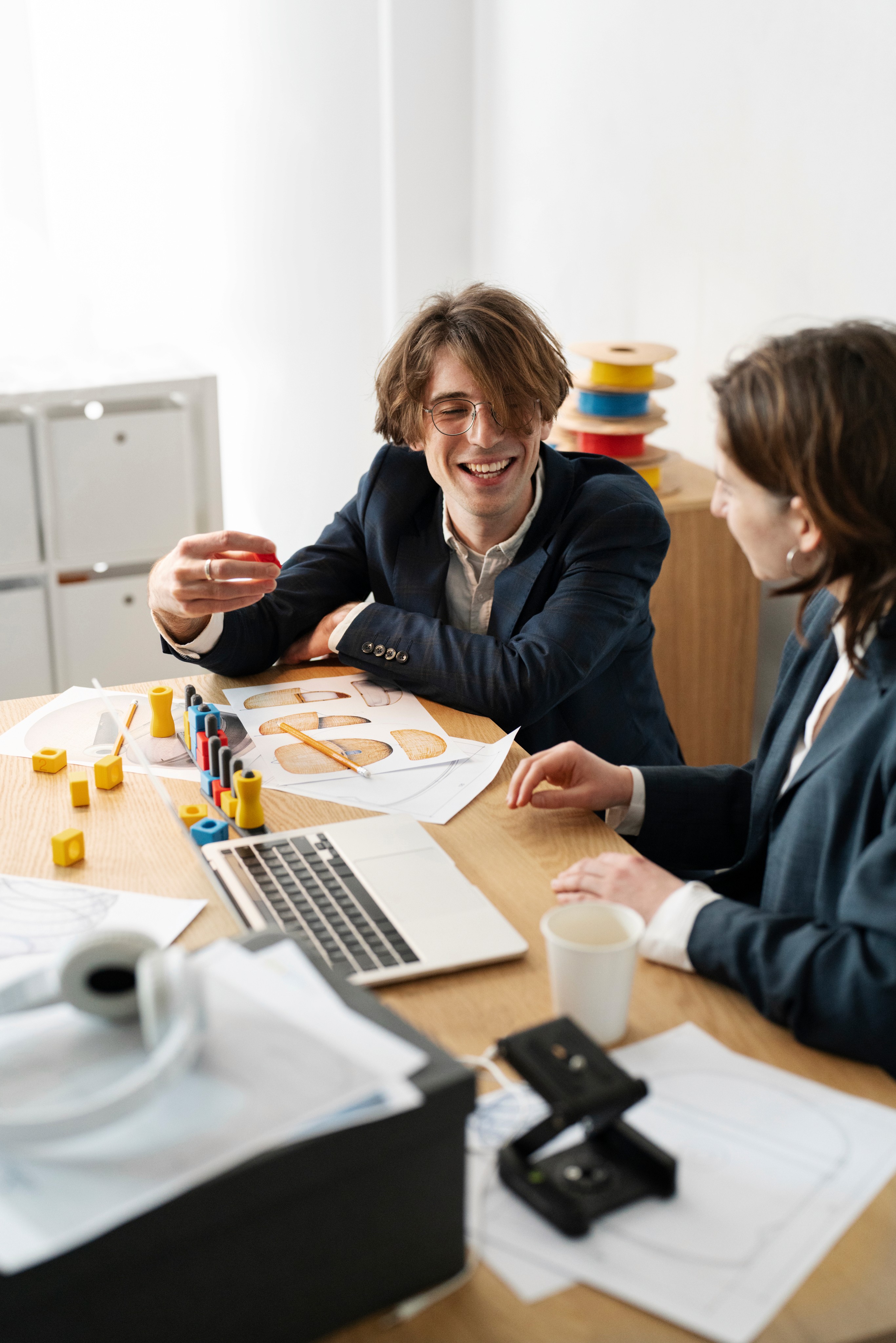 Two people in business attire discussing colorful geometric shapes and drawings at a desk with a laptop.