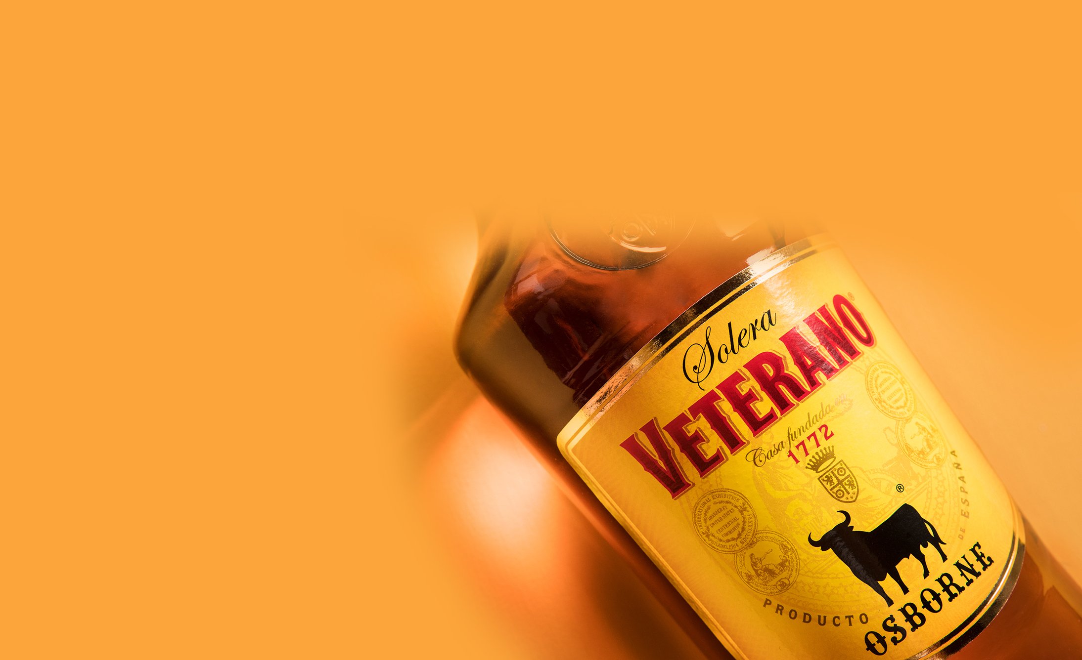 A tilted bottle of Osborne Veterano Solera brandy on an orange background, displaying a bold label with a black bull logo, evoking tradition and warmth.