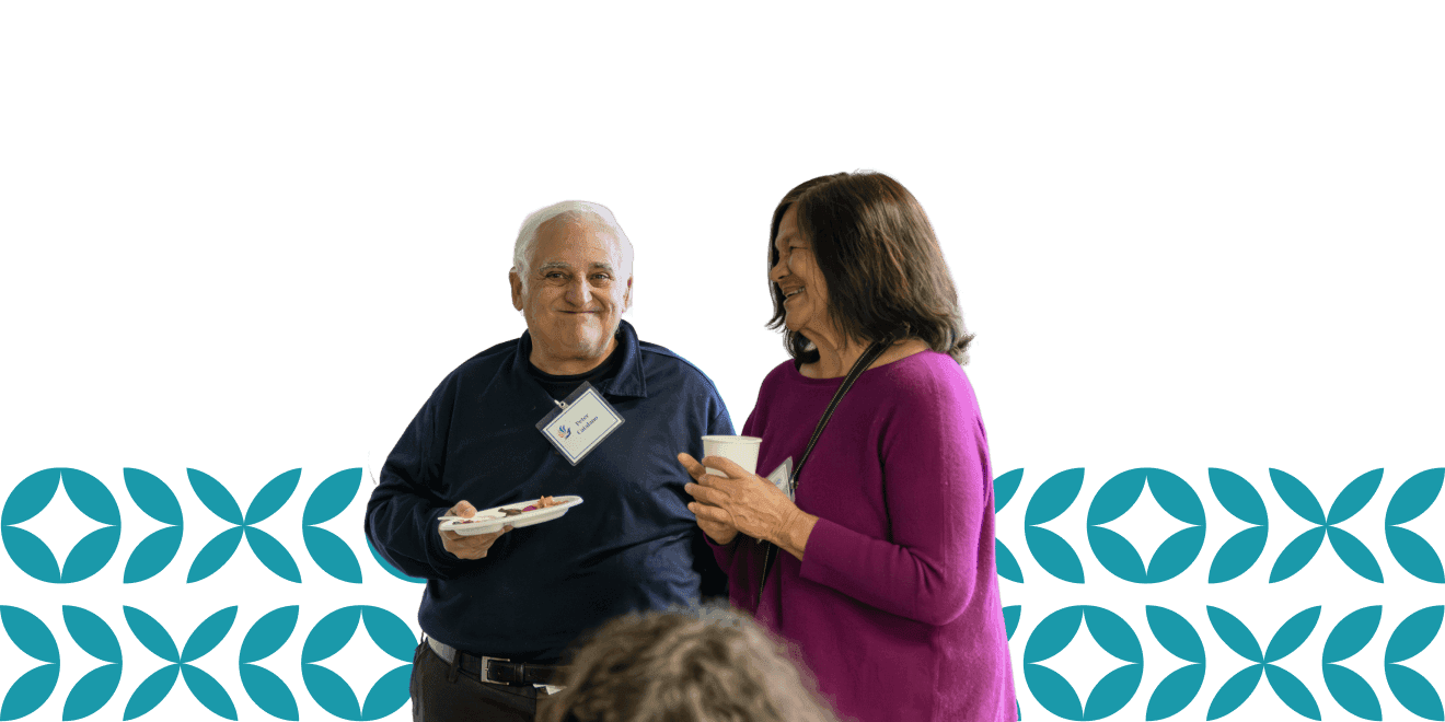 Um homem e uma mulher idosos conversam alegremente enquanto seguram comida e bebidas, em pé diante de um fundo verde-azulado estampado.