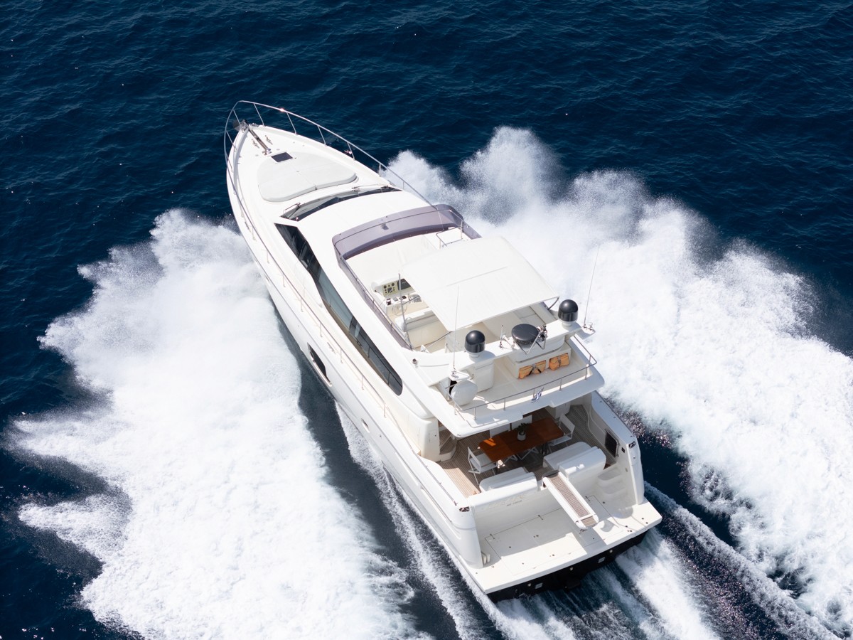White Ferretti 63 motor yacht cruising through deep blue waters with wake trailing behind, blue sky with scattered clouds above.