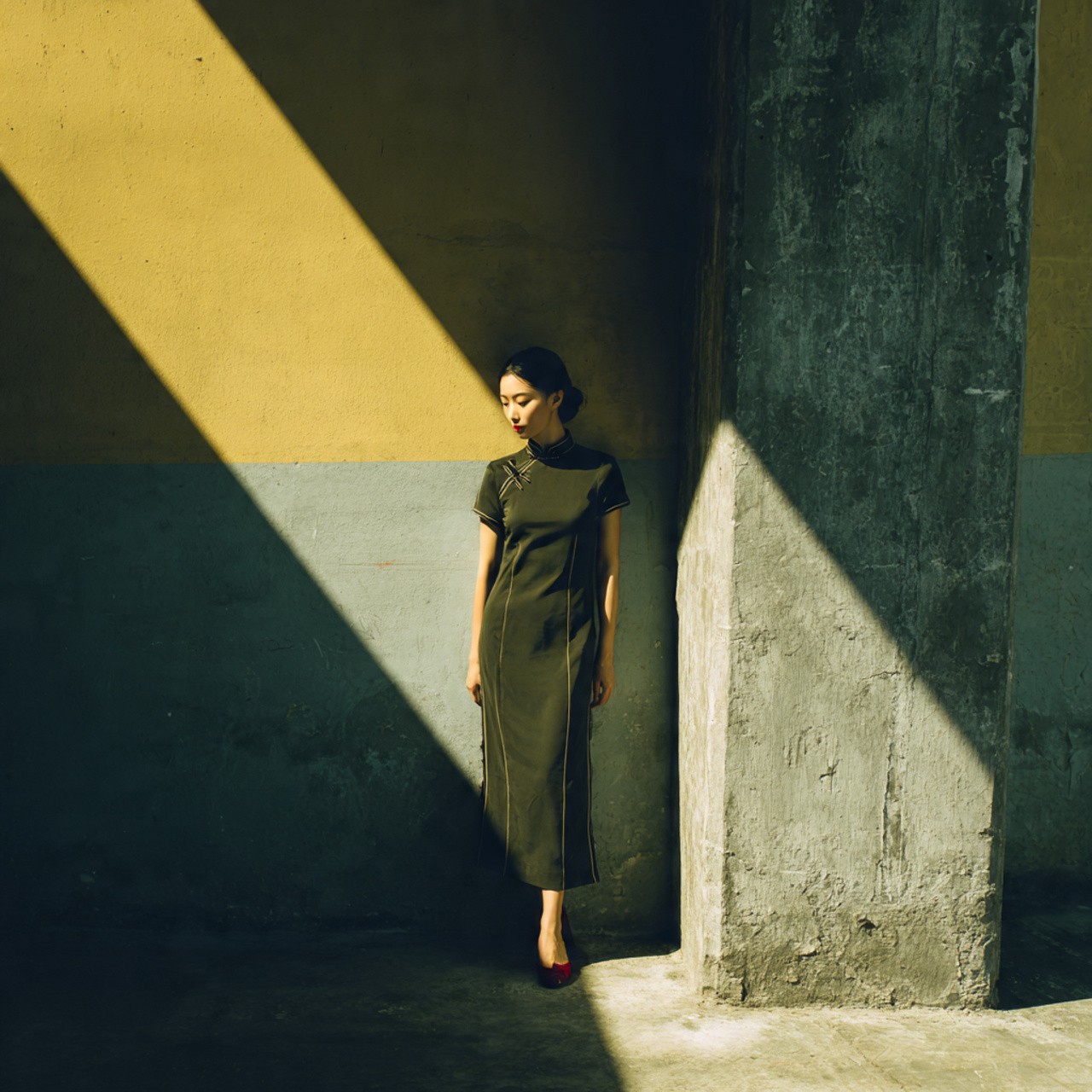 Woman in a dark traditional dress (cheongsam/qipao) standing against a yellow and grey wall in sharp sunlight and shadow.
