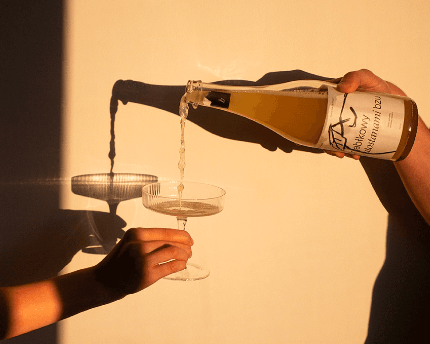 A hand pours a cider from a bottle into a glass, casting a shadow on a light background.