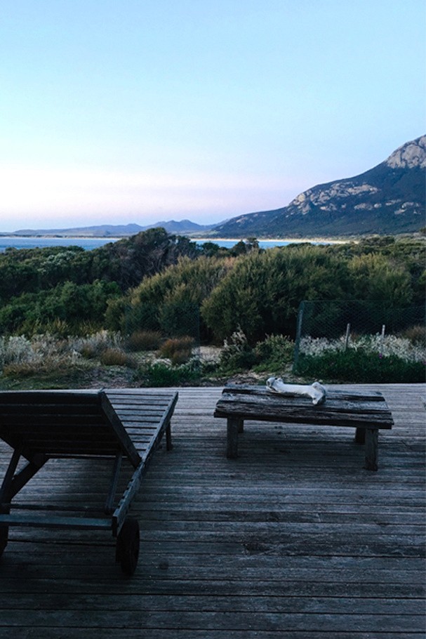A sun lounger and bench on the timber deck at dusk, looking across scrub to the mountains