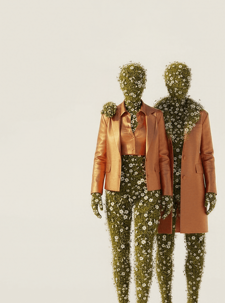 A surrealist digital artwork of three humanoid figures standing side-by-side against a cream background. Each figure is composed of green grass and blooming white daisies where skin would be, including their heads and hands. They are dressed in fashionable, metallic bronze-orange suits and a crop-top blouse. The figure in the center has their arms around the shoulders of the other two, and daisy vines spill over the lapels and collars of their jackets.