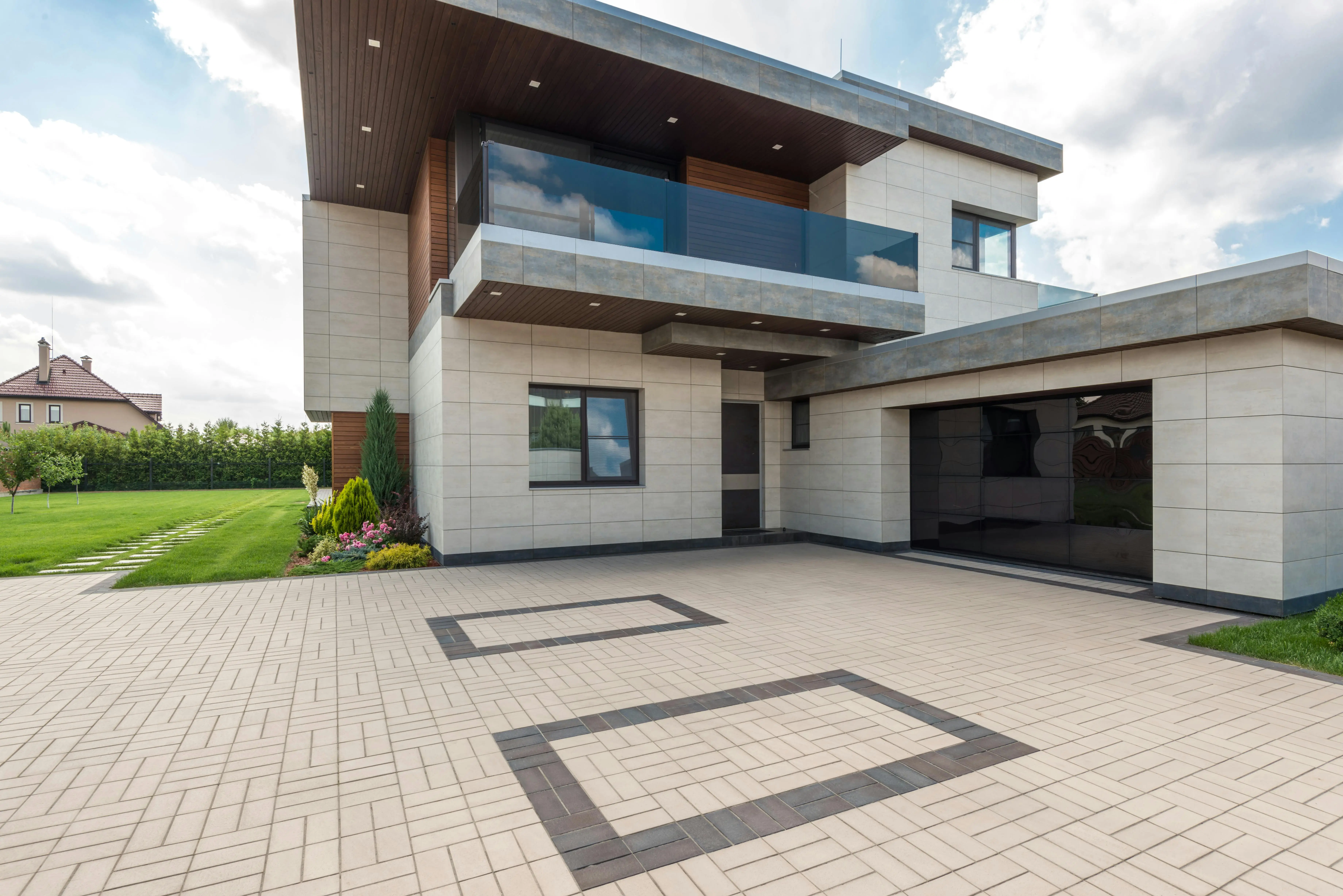 Modern two-story house with sleek design features large windows, a glass balcony, and a spacious paved driveway, surrounded by manicured lawns.