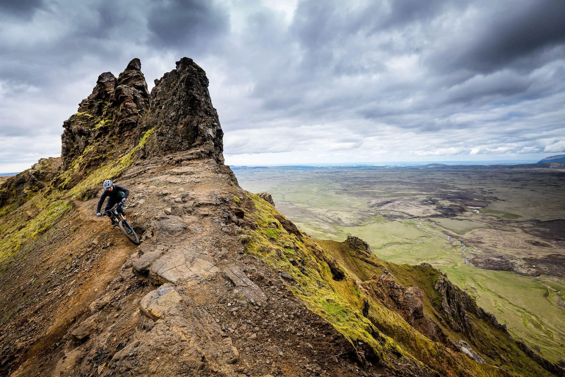 Mountain biker riding a narrow ridge with a wide valley view under heavy clouds.