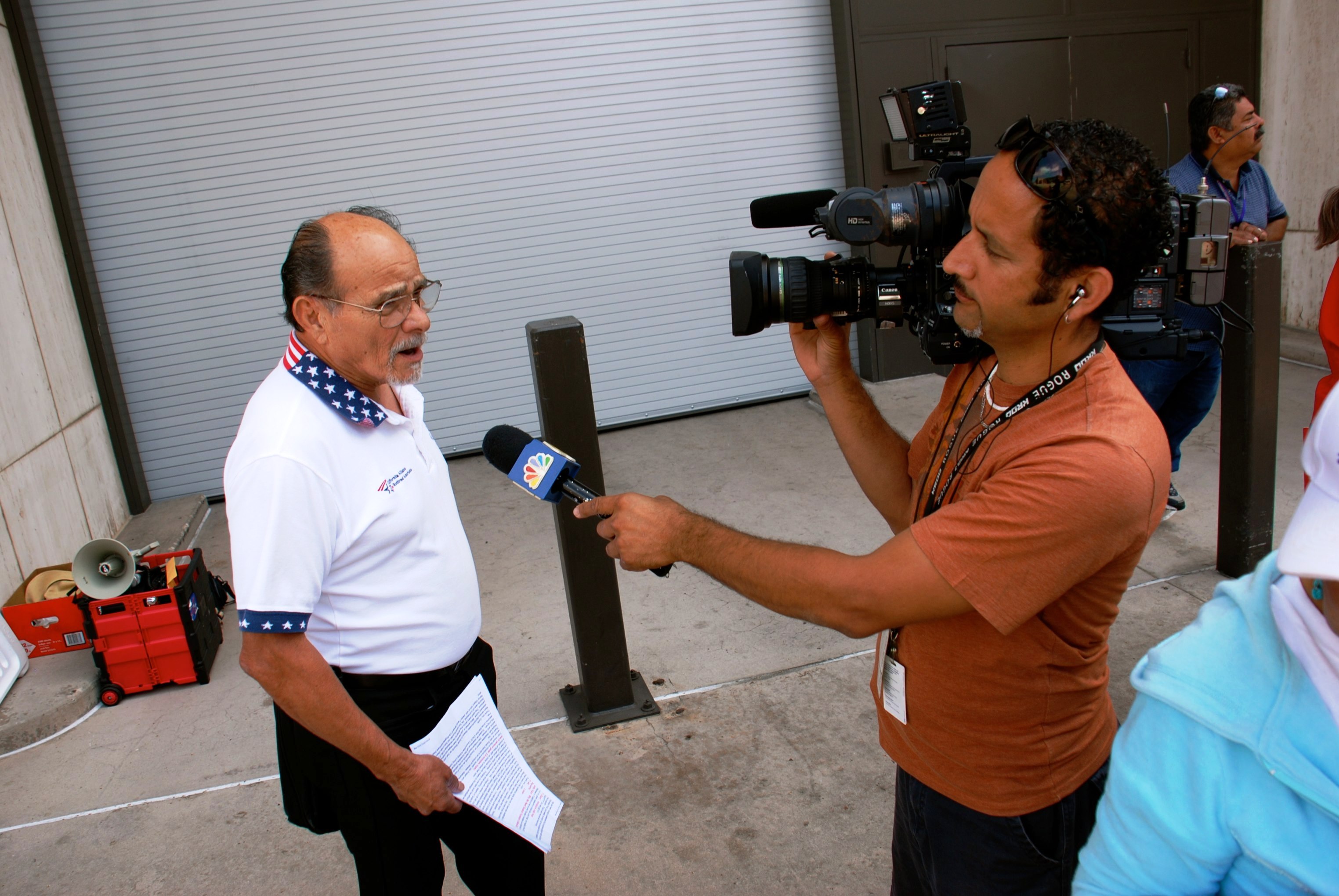 A man holding papers is being interviewed on camera by a news crew outside a building, speaking into a microphone marked with a news station logo.