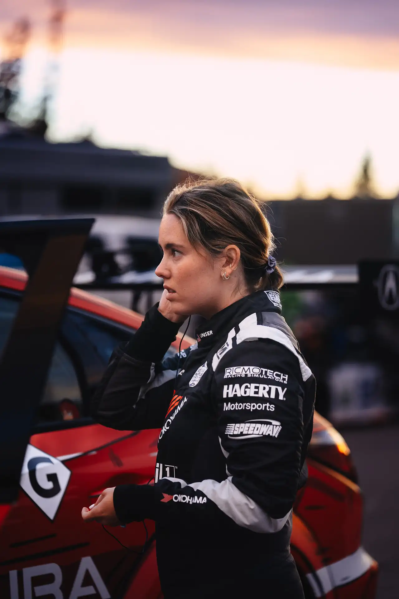 Loni Unser in her driving suit, standing by her red Acura race car, with Hagerty, Ricmotech, and Yokohama logos on her arm.
