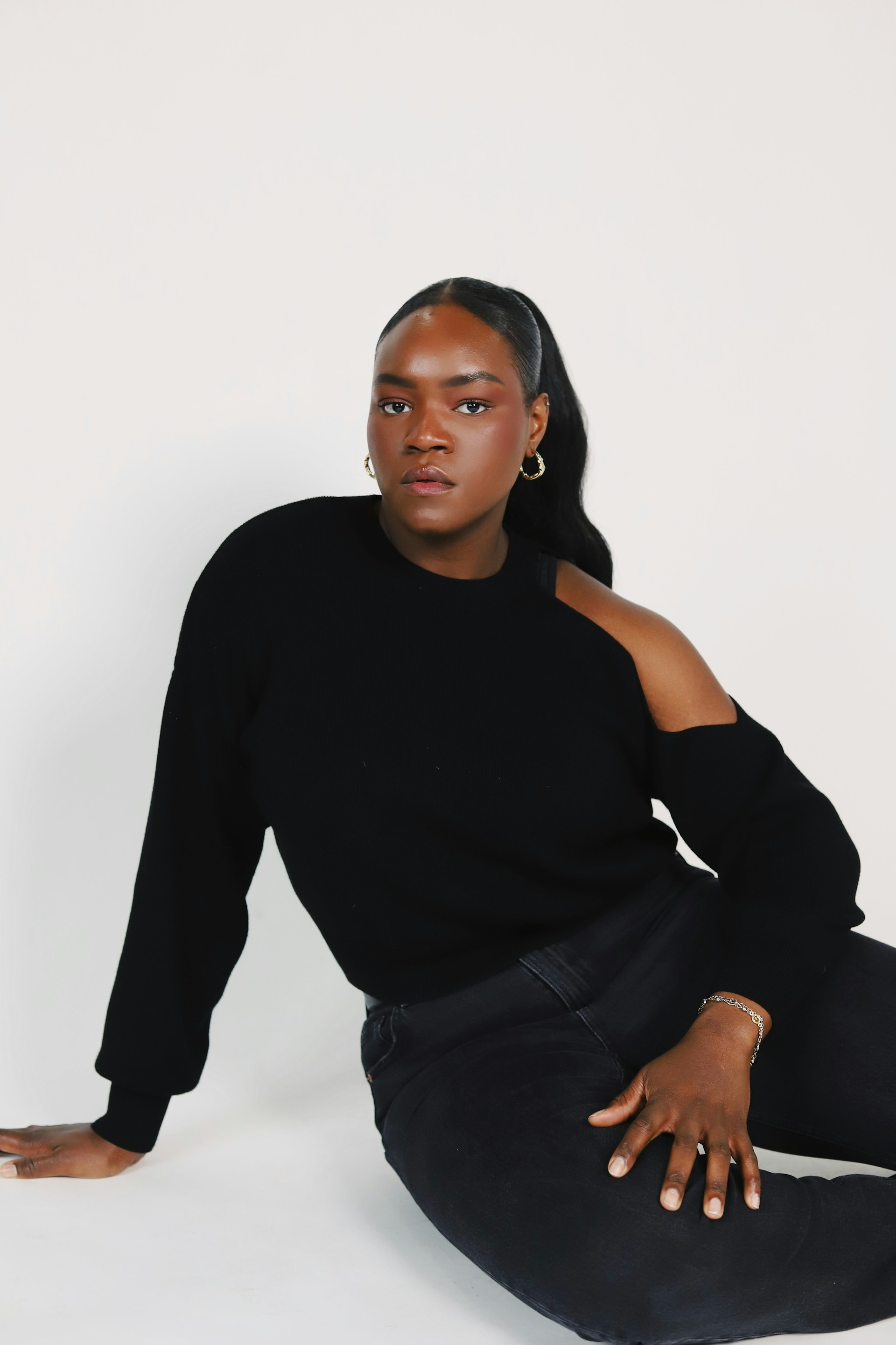A Black woman with a sleek high ponytail poses sitting on the floor against a white background. She is wearing a black cold-shoulder sweater, black jeans, and gold hoop earrings.