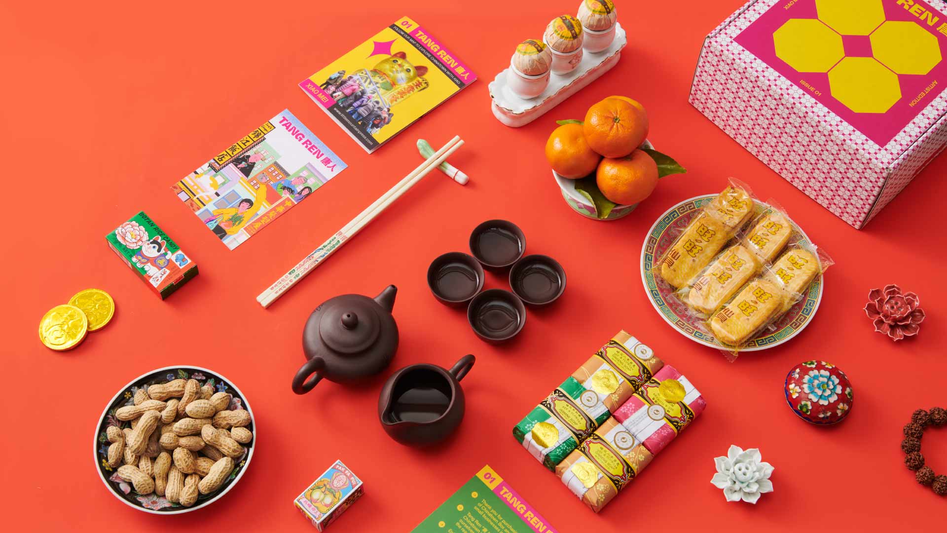 The image shows a collection of items arranged on a vibrant red background. These items include a black teapot, two black teacups, a white plate with orange slices, a box of cookies, a small decorative flower, a small red box, a yellow booklet, a pair of chopsticks, a small white stick, a small red candle holder with candles, a small box of matches, a small green box, a white bowl with peanuts, a small red bowl with decorative elements, and a bracelet made of dark beads. The items are mostly in close proximity to each other, creating a visually interesting arrangement on the red surface.