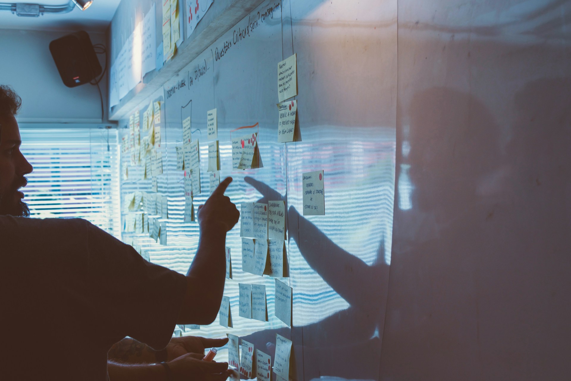 A man stands in front of a whiteboard covered in sticky notes, pointing to one of them. Another person's hand is visible in the foreground, also pointing at the board.