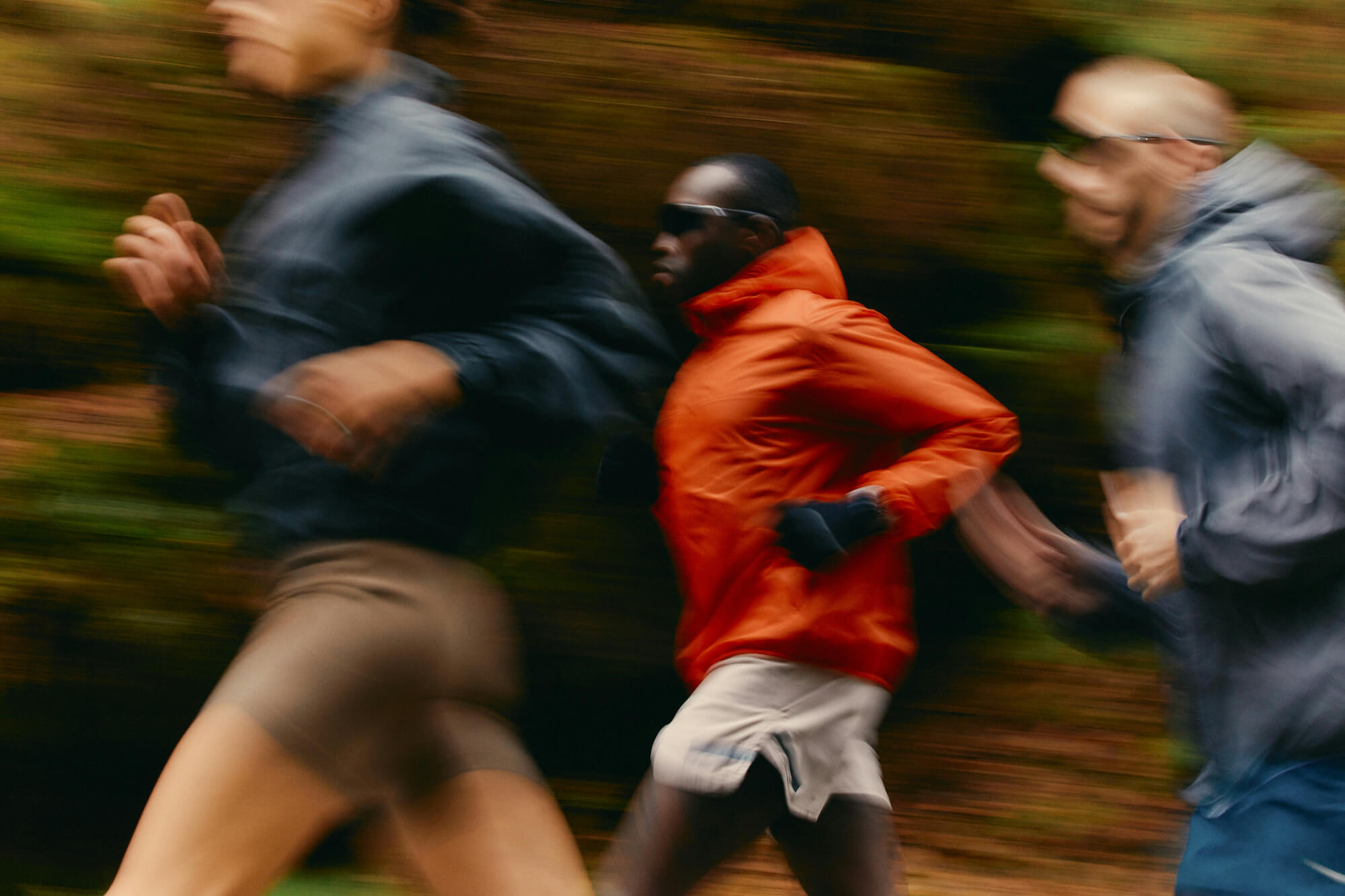 Three people running in motion blur; one wears an orange jacket, others in blue. They convey speed and focus against a blurred forest background.