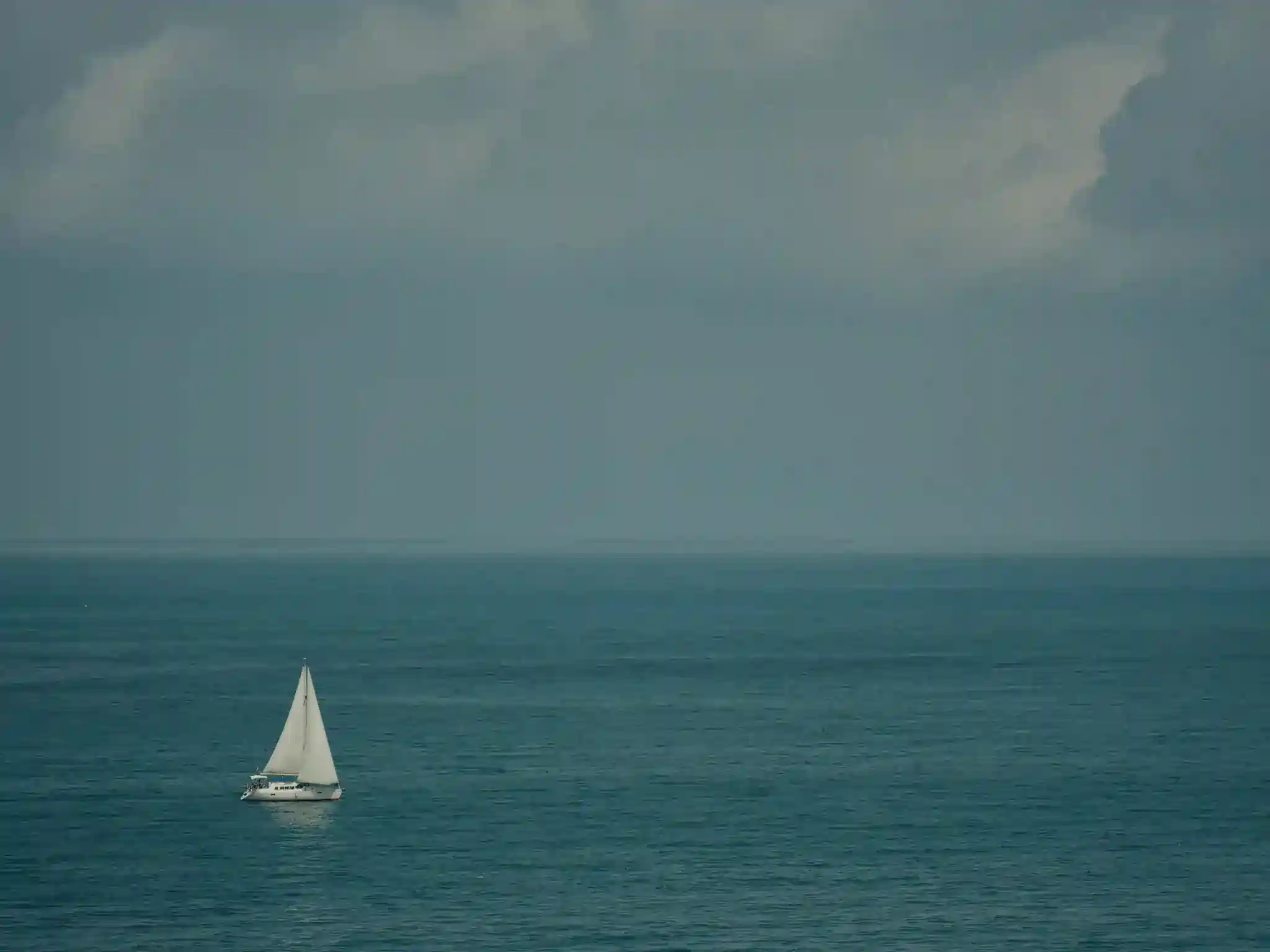  single white sailboat positioned in the lower left portion of the frame. The sailboat is peacefully gliding across a vast expanse of calm, teal-blue water. The horizon line is almost imperceptible, blending smoothly into the overcast sky above.