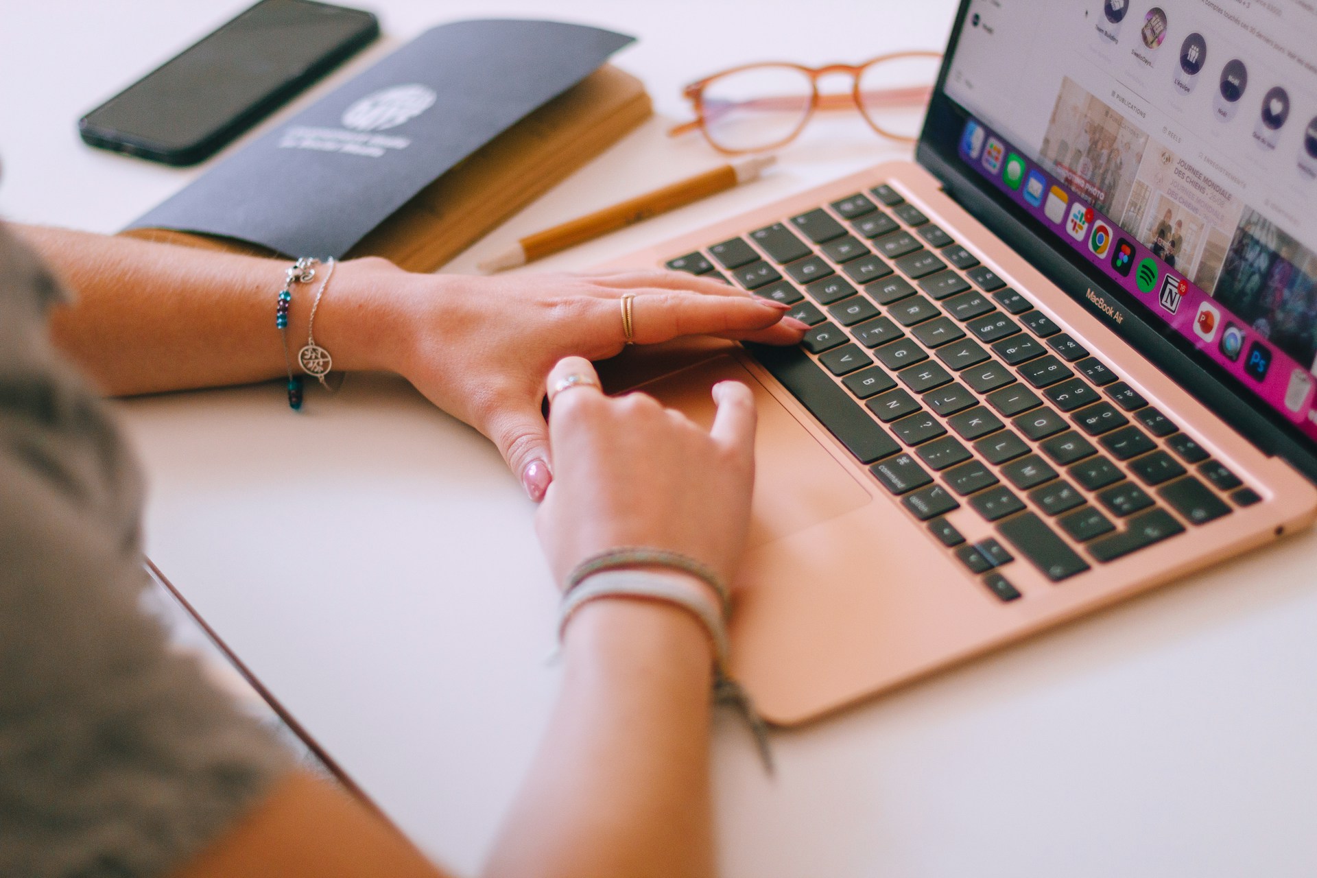 A person's hands are typing on the keyboard of a gold or rose-gold colored laptop, with a smartphone, glasses, and a folder nearby on a white desk.