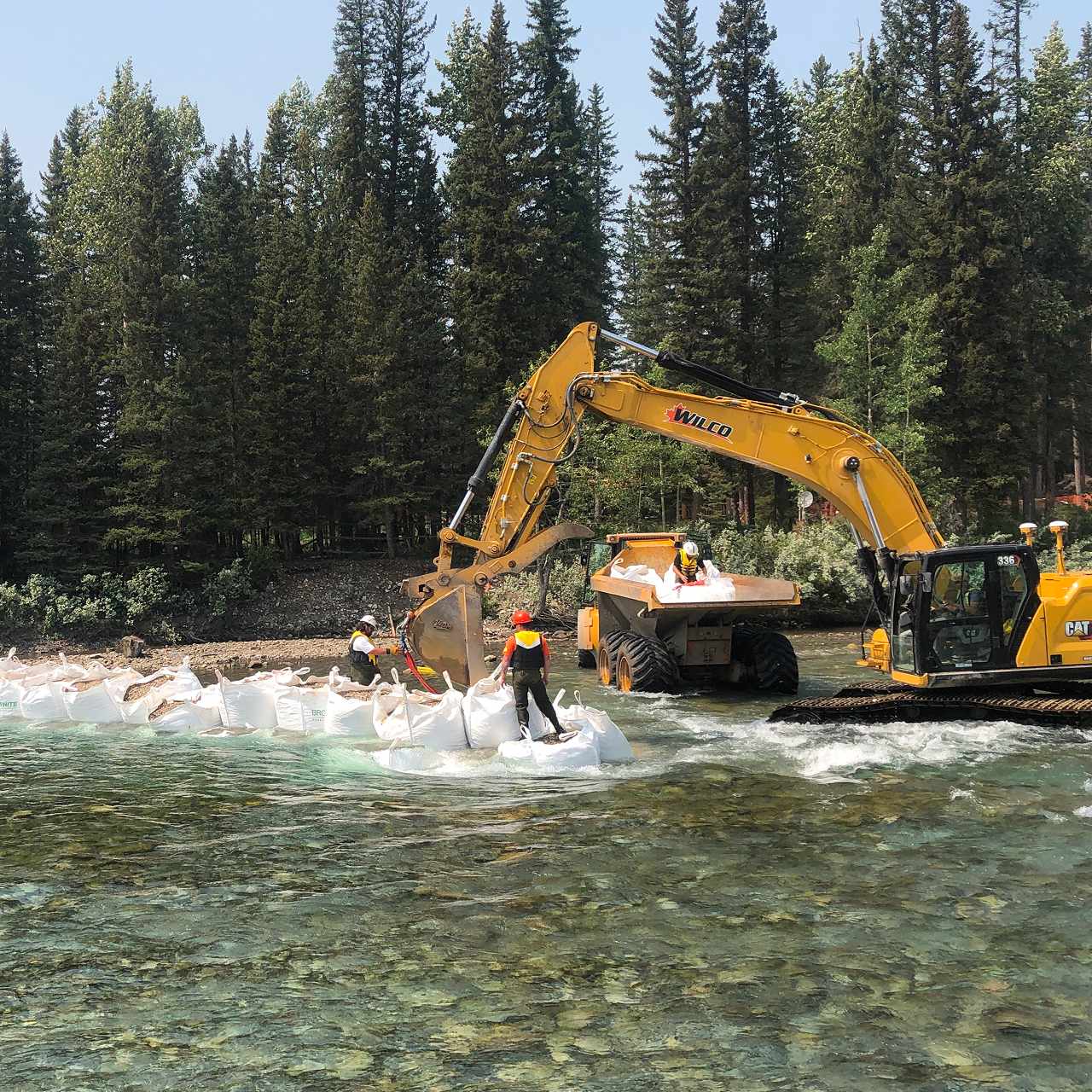 Construction access road and retaining wall excavation at Cougar Creek Dam site in Canmore with heavy equipment working among snow-dusted Rocky Mountain peaks and forest