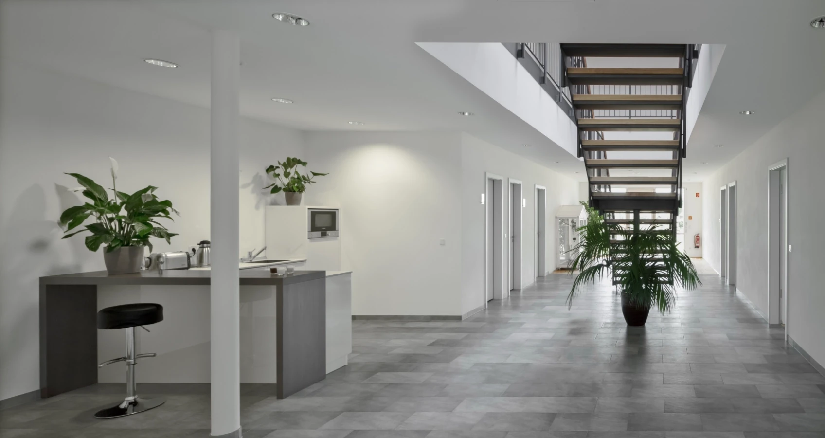 Moderner Flur mit offener Treppe, Küchenzeile und Pflanzen, grauer Boden und weiße Wände.