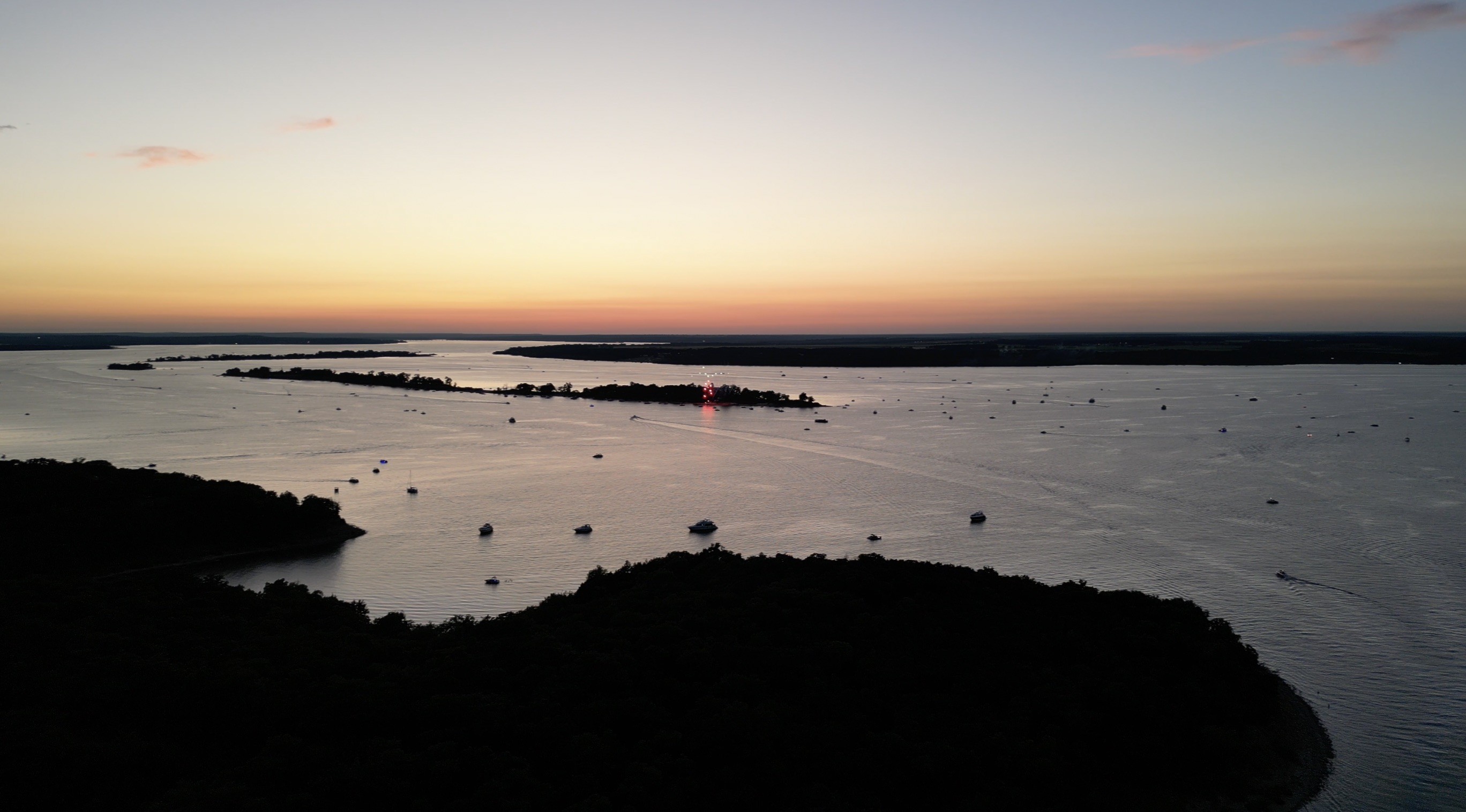 A serene aerial view at sunset captures a wide, calm river dotted with numerous boats, with the sky transitioning from soft orange to darker shades, and a tree-lined island in the background.
