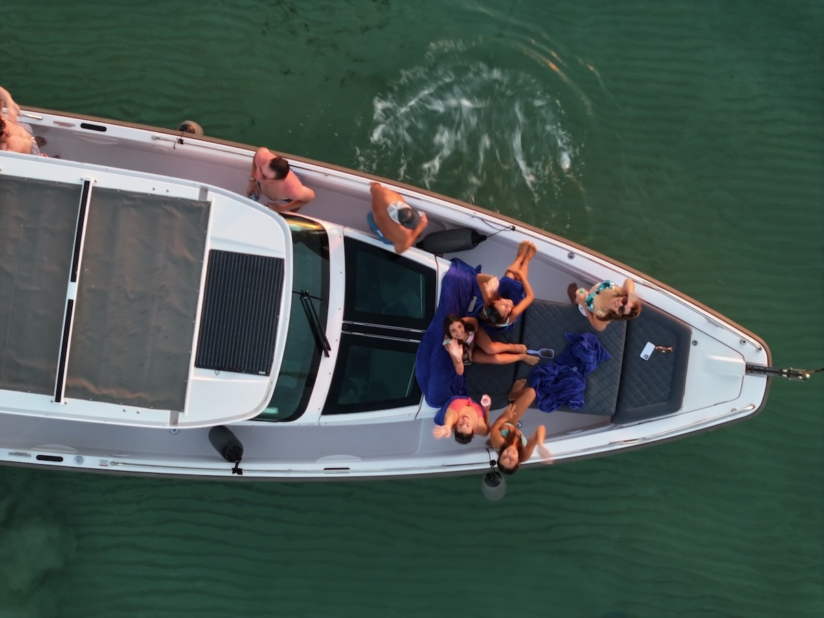 Aerial view of white Axopar 37 motor yacht with guests relaxing on blue cushioned seating in crystal-clear turquoise waters.