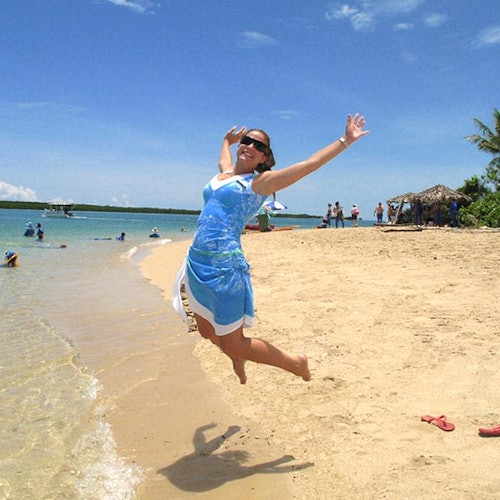 ผู้หญิงในชุดเดรสสีน้ำเงินกระโดดอย่างร่าเริงบนชายหาดที่เต็มไปด้วยทราย โดยมีผู้คน กระท่อม และท้องฟ้าสีฟ้าใสอยู่ด้านหลัง