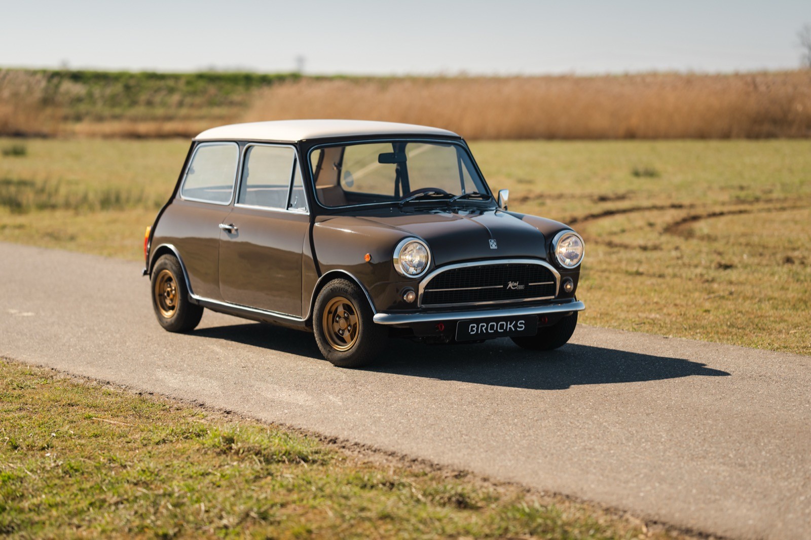 A classic small car parked on a rural road, surrounded by grassy fields and a clear sky.