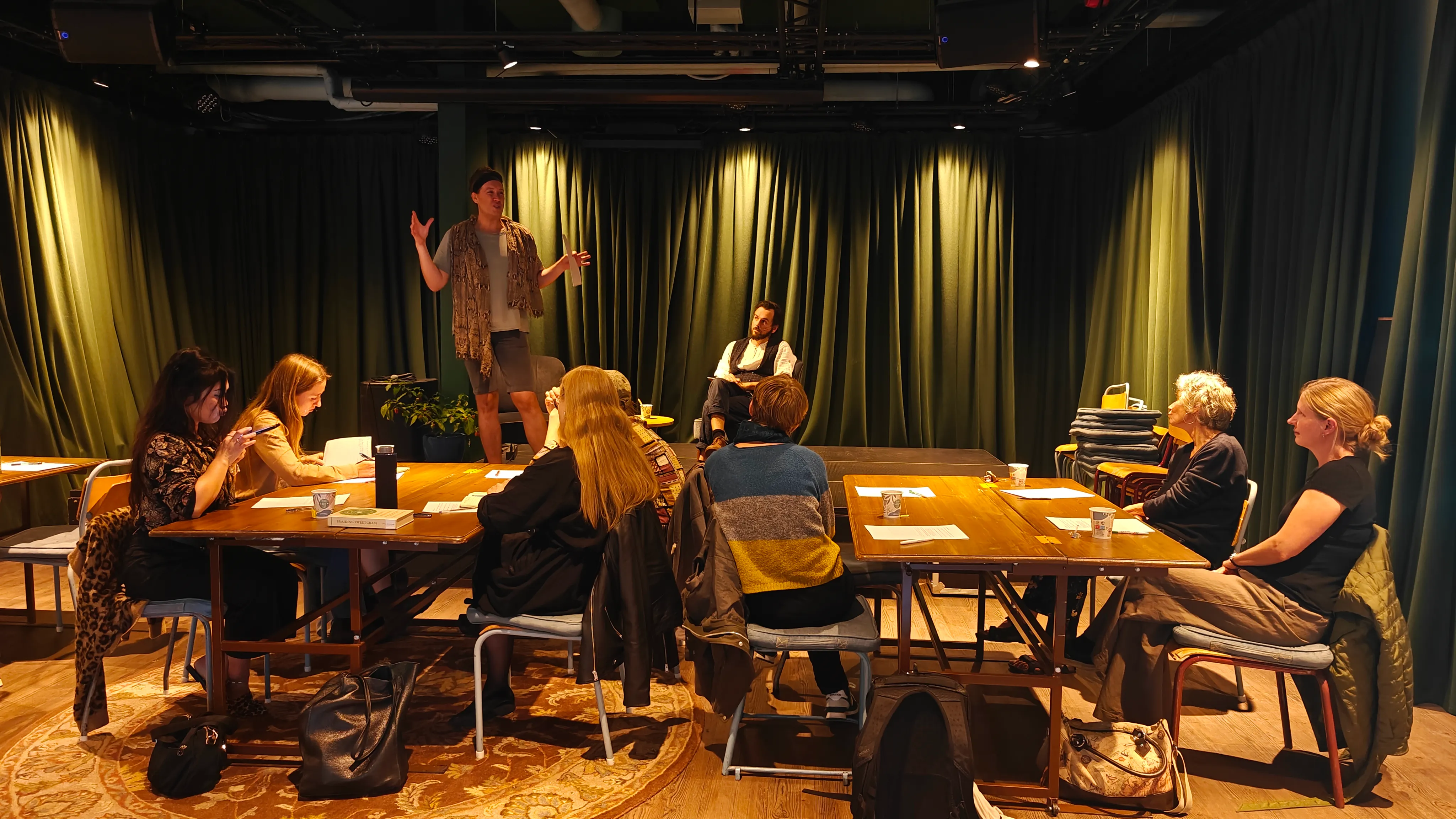 Several groups of people sitting down around tables while two people are on the stage talking