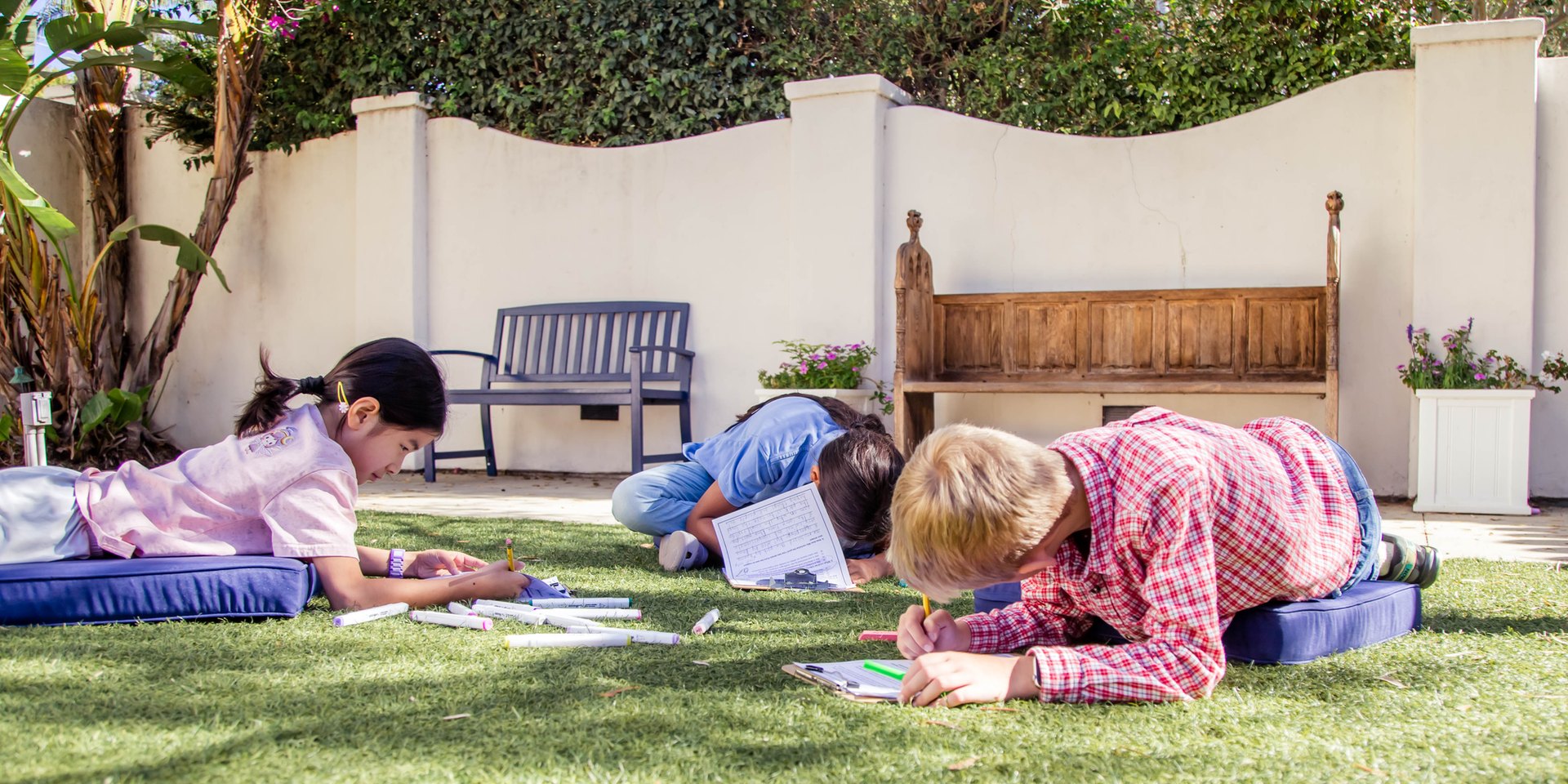Three students lying on cushions on the lawn, writing in a walled garden courtyard