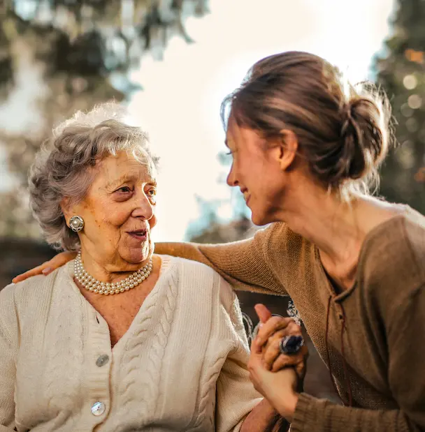 A younger woman leaning in warmly toward an older woman, holding her hand and sharing a gentle, connected moment together outdoors.