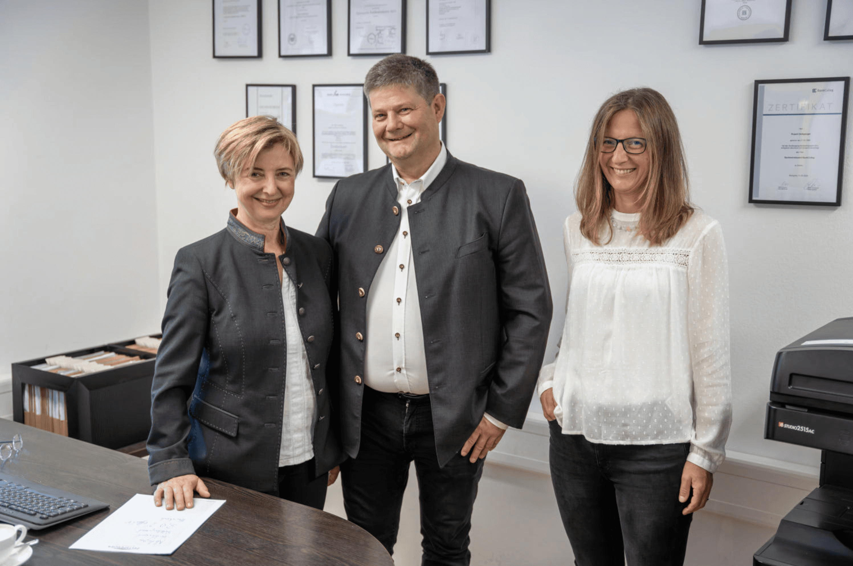 Hubert und Hildegard Zeltsperger stehen gemeinsam mit Alexandra Zierer in ihrem Büro mit Zertifikaten an der Wand