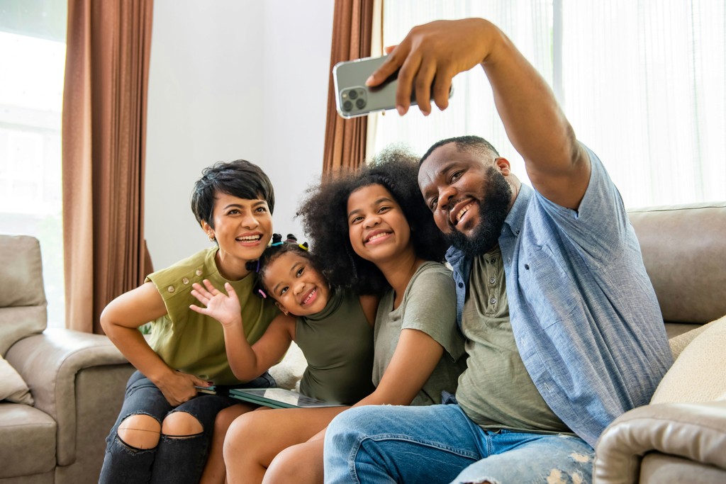 A happy family of four takes a cheerful selfie together in their living room. The parents and two daughters, all smiling and waving, gather closely on a comfortable couch. The scene captures the warmth and joy of family bonding and the delight of capturing moments together with a smartphone.