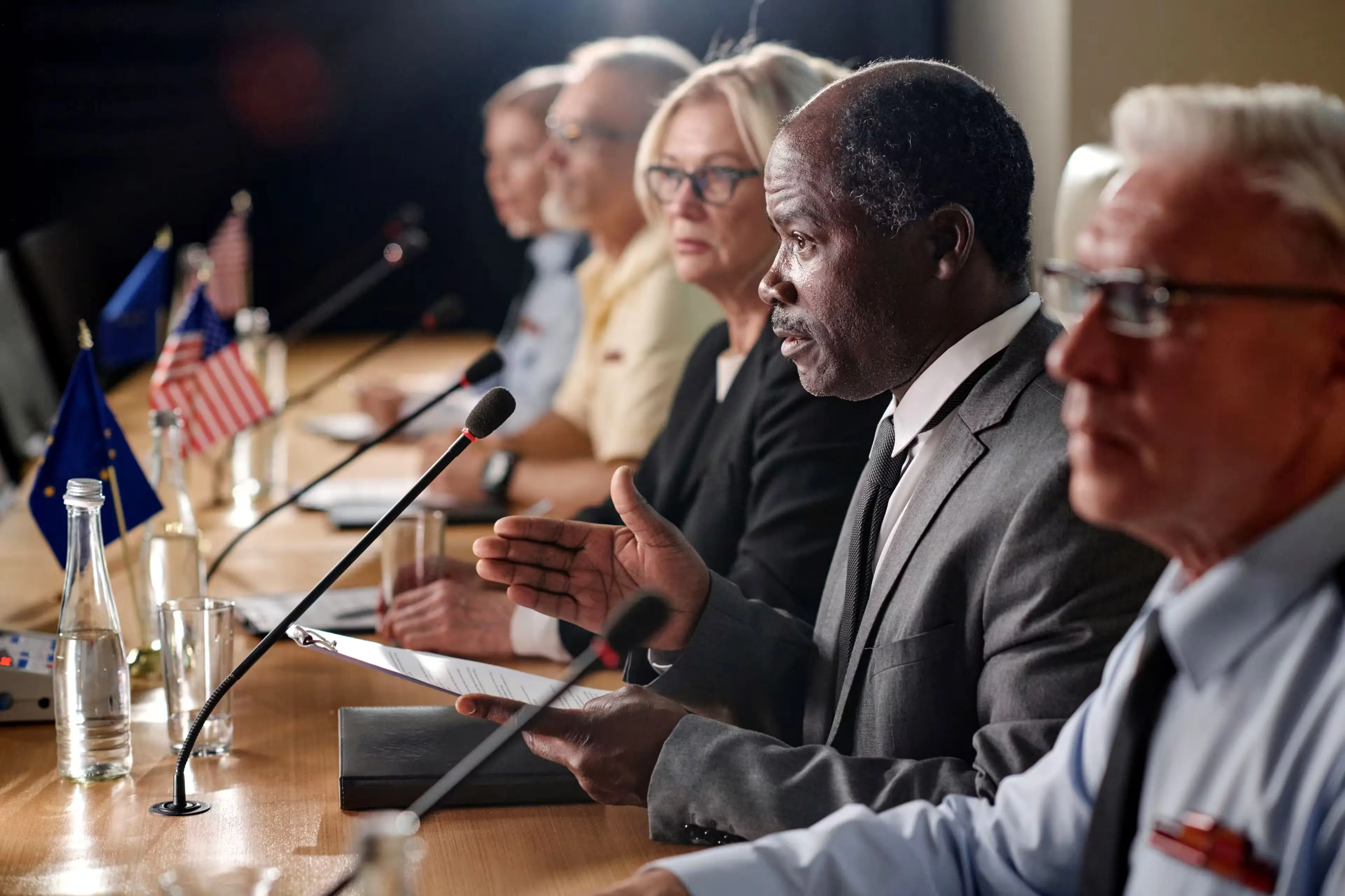 Panel of diverse professionals speaking at a conference table equipped with microphones, with small national flags and glass bottles on display.