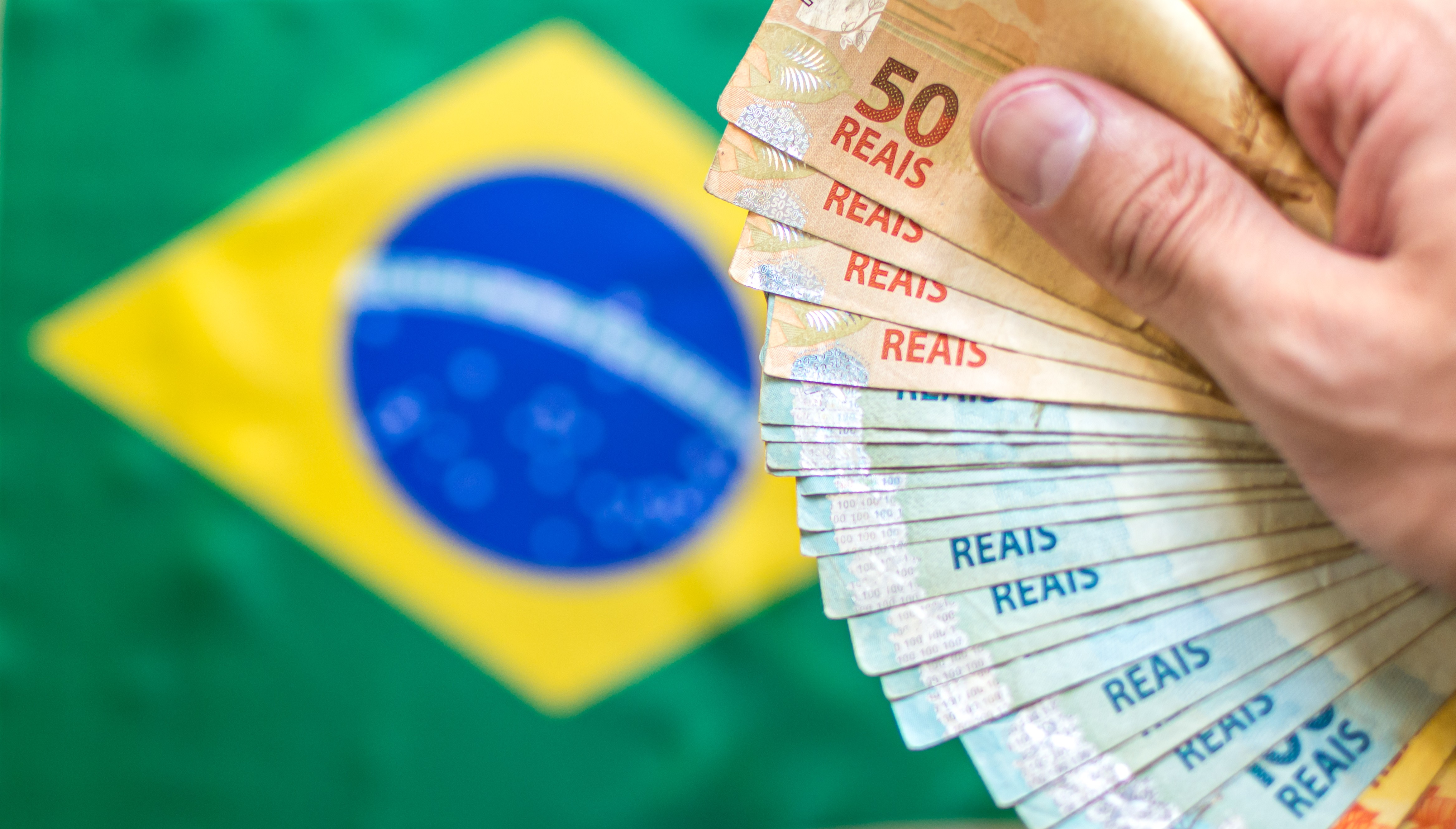 Hand holding Brazilian real banknotes in front of a blurred Brazilian flag.