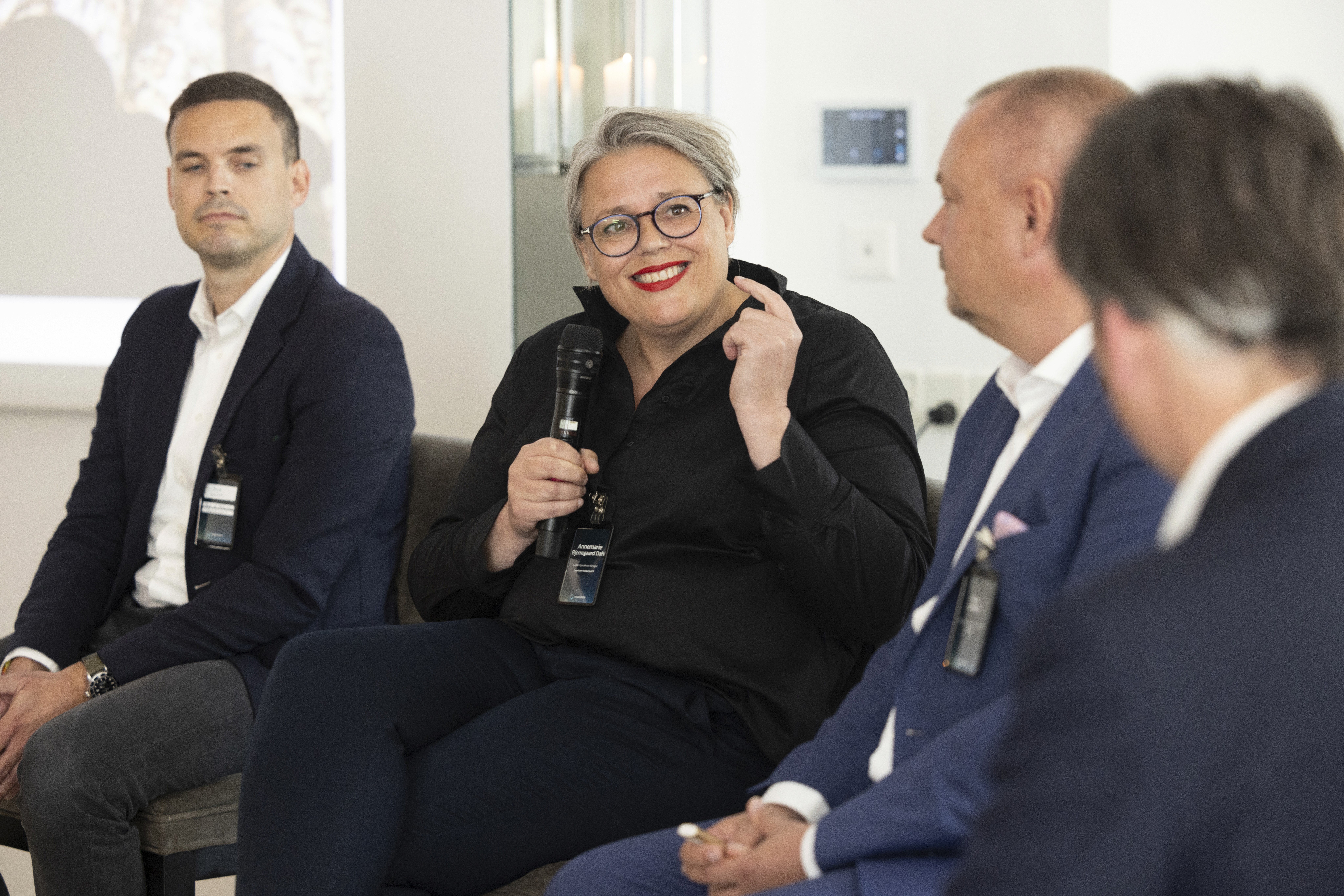 Smiling woman speaking into a handheld microphone on a panel with three men in suit jackets