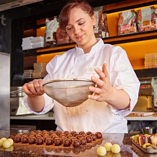 在厨房里,一位身穿白色厨师制服的厨师在多排巧克力松露上撒着可可粉。背景中有放置各种配料的架子。