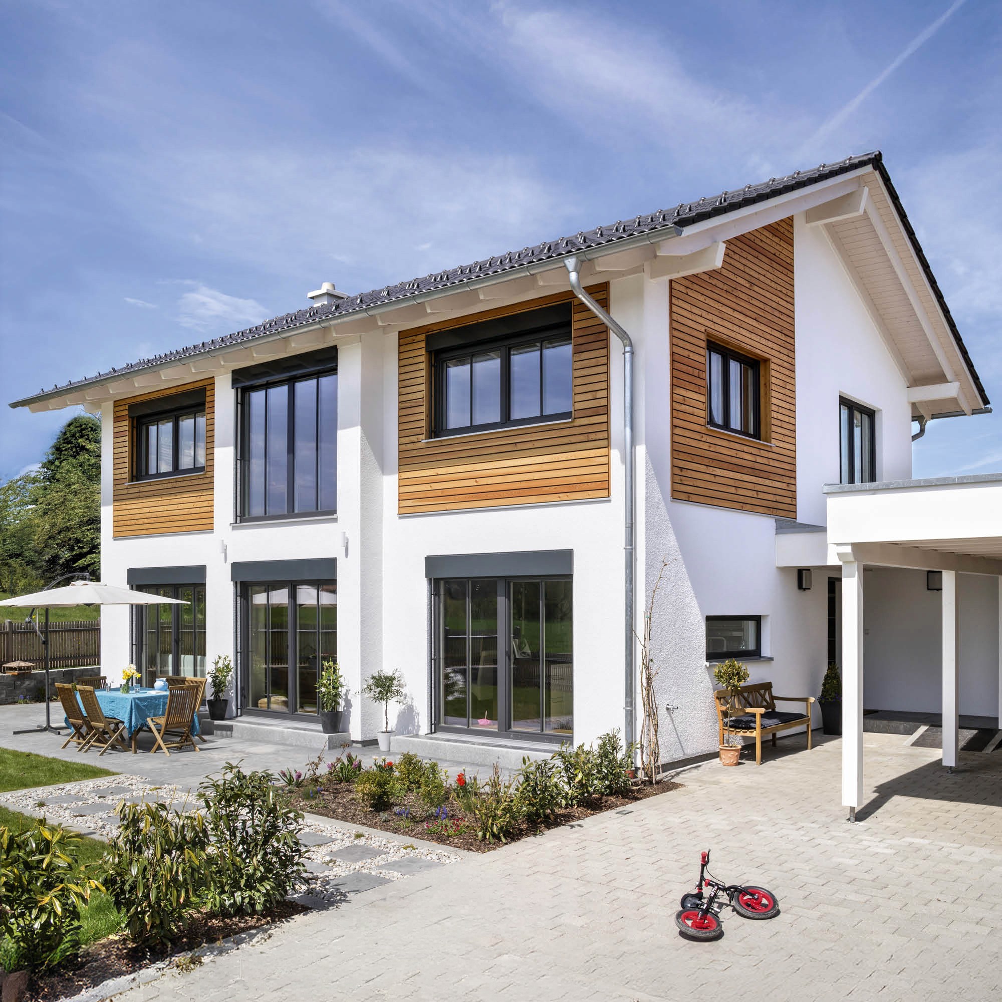 Modernes Einfamilienhaus mit weißer Fassade und Holzverkleidung, großzügigen Fenstern, Carport und Garten.