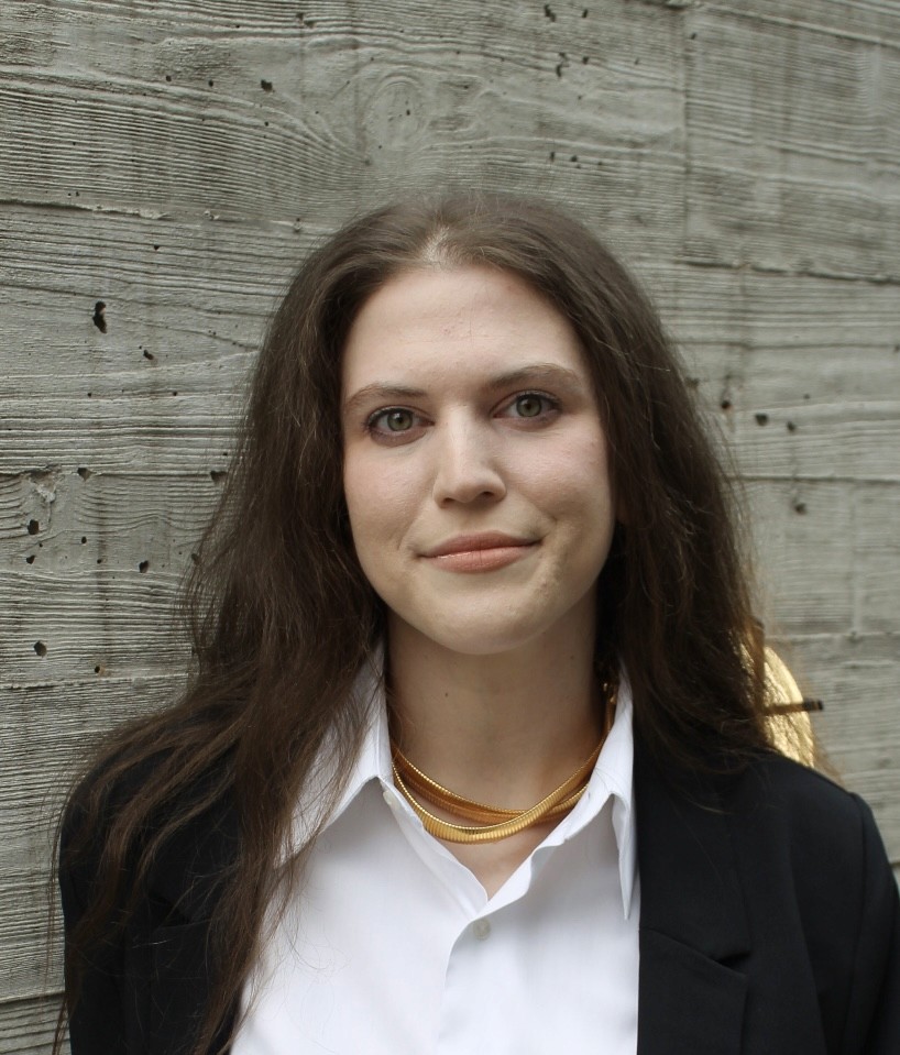 Founder Miriam Tenner is in front of of a cement background that has a wood grain impression finish. She sports auburn center parted long hair, has a white button down, black blazer and a vintage gold jewelry necklce.