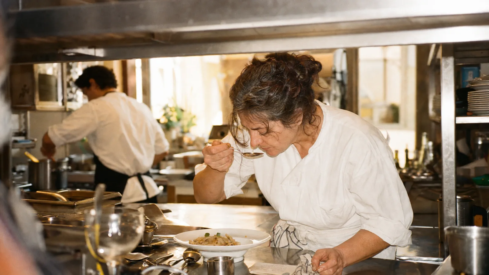 Cuoca assaggia con il cucchiaio un piatto di prova al passe accanto alla scheda ricetta