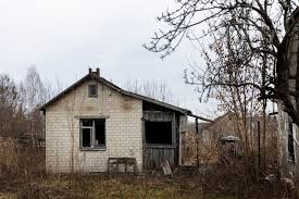 A small, derelict single-story rural house with white brick walls under an overcast sky. The house has a steeply pitched roof, one broken or missing window pane, and a dark, dilapidated entryway on the right. The foreground is overgrown with dry, brown weeds and tall, dead grasses, and the bare branches of a tree frame the upper right corner.