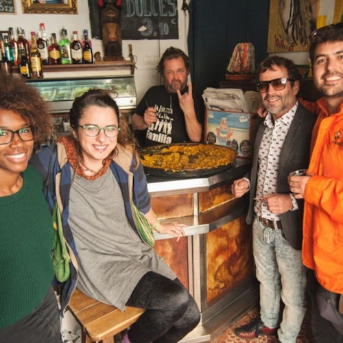 四个人在酒吧里微笑着摆姿势，靠近一个巨大的西班牙海鲜饭锅。柜台后面的一名男子做出了摇滚手势。