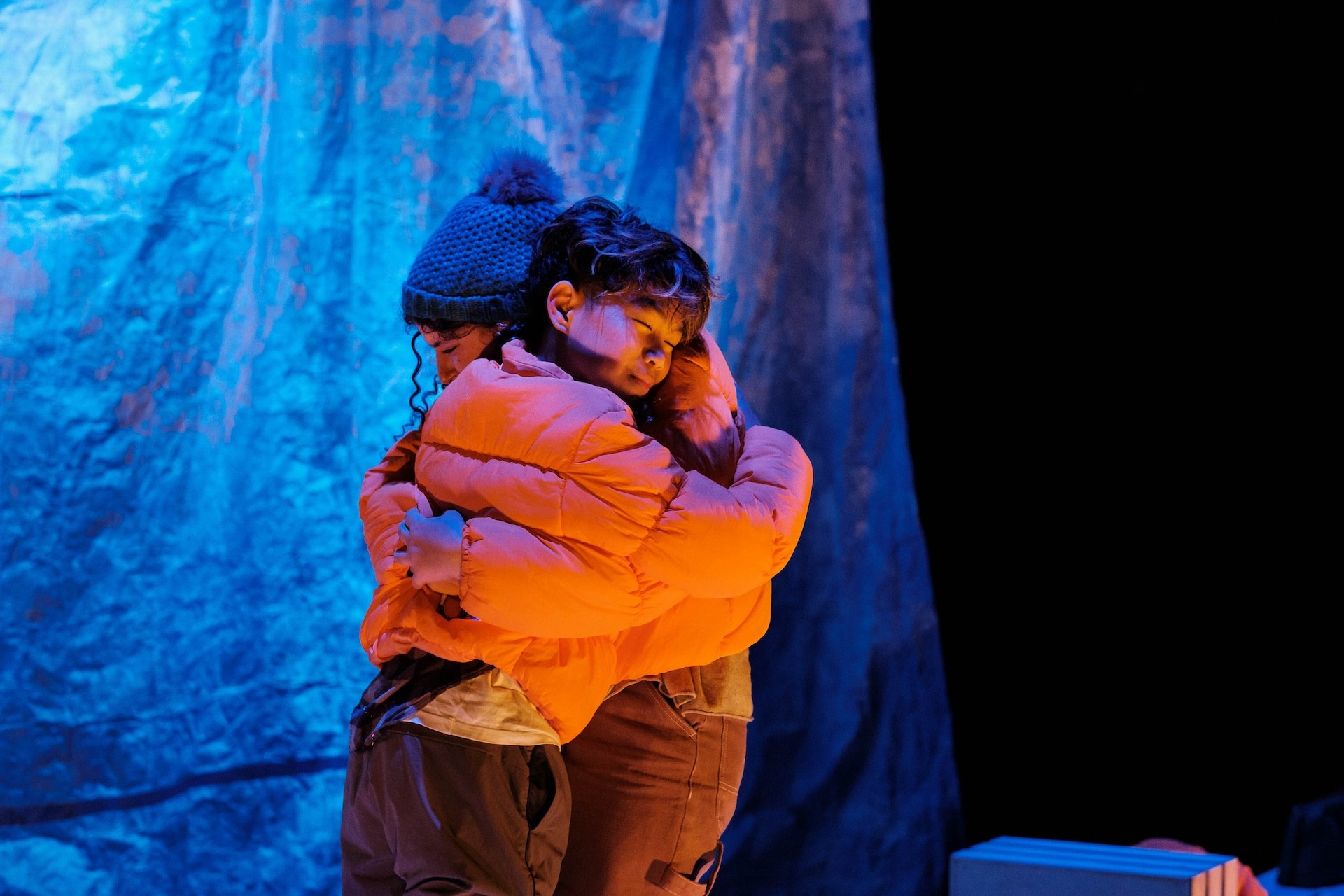 Two performers in orange puffer jackets share a tight embrace. Their eyes are closed, and they stand against a cool blue backdrop that suggests an icy or outdoor environment.